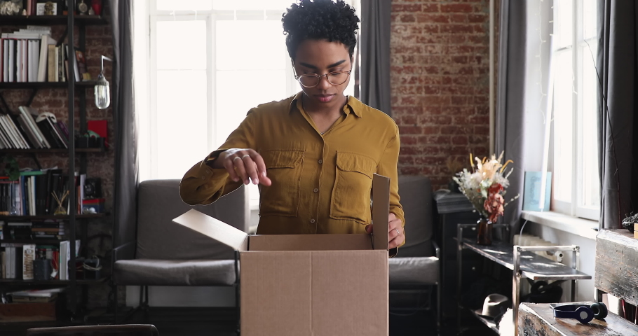 Curious smiling Black gen z female unpack postal parcel open big box with consumer goods purchased on sale at ecommerce web store. Happy millennial female client receive unbox mail delivery package Royalty-Free Stock Footage #1089117349