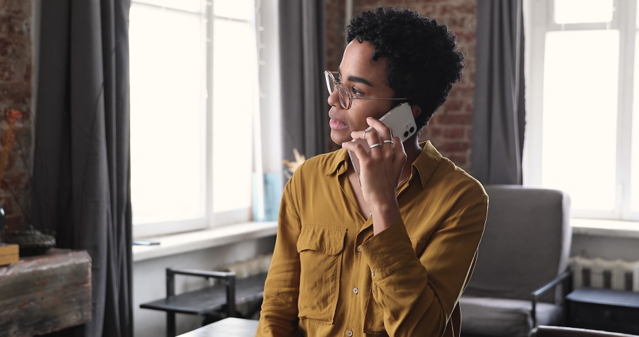Pleasant young African American lady chat with friend using mobile phone listen to news answer call holding device close at ear. Happy relaxed teen female in eyeglasses talk speak on telephone indoors Royalty-Free Stock Footage #1089117363