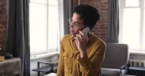 Pleasant young African American lady chat with friend using mobile phone listen to news answer call holding device close at ear. Happy relaxed teen female in eyeglasses talk speak on telephone indoors