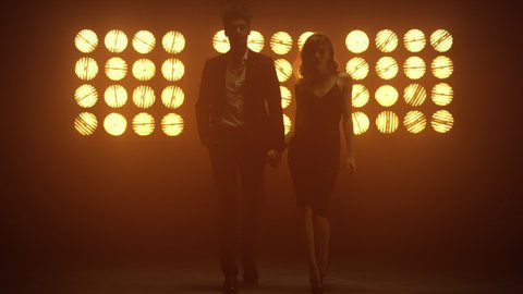 Sexy couple walking together confidently holding hands on dance stage. Young pair posing standing in front club lights. Handsome man wearing suit moving forward with hot woman in elegant dress indoors