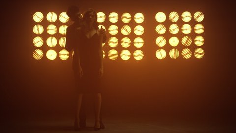 Sexy man caressing gorgeous woman relaxing in night club. Attractive guy standing behind beautiful girl touching skin in spotlights. Passionate young pair walking together at show stage. Love concept