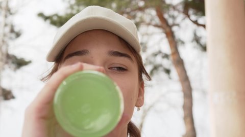 a girl in a cap drinks a green carbonated drink from a glass bottle and eats flatbread. fast food and a snack in nature on a walk in the park or grove.
