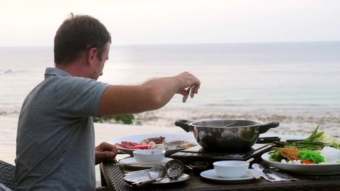 Sukiyaki or Shabu shabu. Smiling inspired man cooking fresh seafood and meat in hot pot in Japanese restaurant with sea view. Happy caucasian man eating asian food