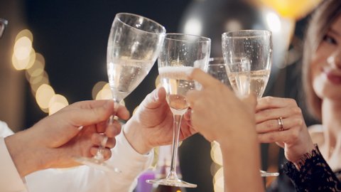 Portraits of young multiracial girls sitting at festive table and celebrating holiday. Women congratulating friends, smiling, laughing, blowing whistle, clinking glasses of champagne and drinking wine.