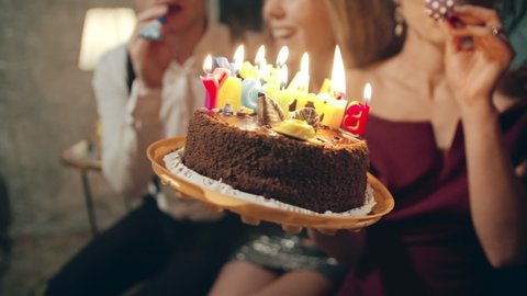 Portrait of asian girl wearing birthday or party cap, blowing whistle and holding birthday cake with burning candles donated by caucasian friends. Happy millennials spending time together at party.