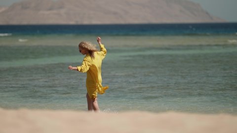 a curly-haired blonde in sunglasses and a yellow fluttering dress walks on a shallow sea and wets hands. the general plan