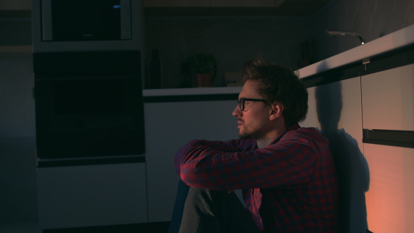 Young man with a thoughtful look sits on the kitchen floor and looks ahead, illuminated by the light of the sunset. Pensive, sad, immersed in looking away thoughts dreams | Shutterstock HD Video #1089123407
