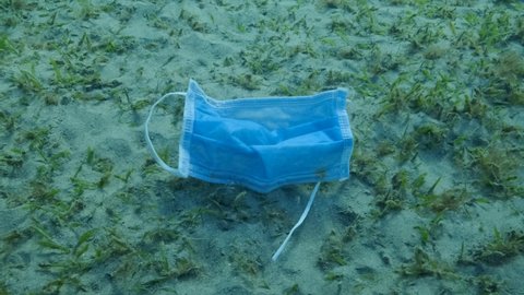 Slow motion, Used face mask lies on sandy bottom covered with green seagrass in sunlight. Discarded face masks polluting seabed since Coronavirus COVID-19. Pollution of Oceans. Camera moves forward