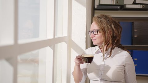 Successful business woman enjoys drinking coffee and looking out window on sunny morning in office, feeling happiness.