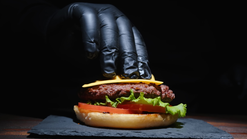 Chef making tasty burger on serving plate on black background close-up. Cheeseburger with beef patty, cheese, tomato, onion, fried bun, lettuce, sauce.