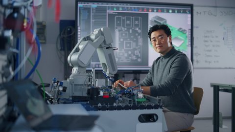 Asian Male Engineer Sitting at Table Using Robot Hand and Engineering; Turning and Looking at Camera. Robot is Moving Under his Control. Education and Robotics Concept