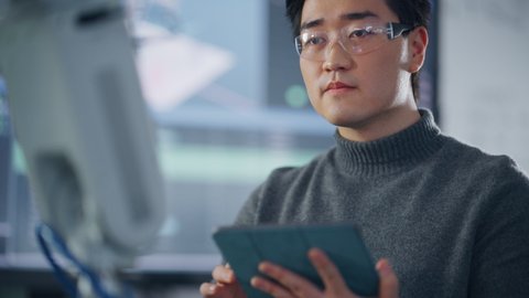 Male Student Typing at Tablet while Programming Bionic Claw At Factory. Robot is Moving Under his Control. Young Developer in Robotics Facility Working on Software Production. Close Up View