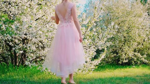 Young happy woman rear view, evening festive hairstyle, long hair gathered up cute smiling face. Girl trendy graduate. Pink bridal dress open back. Hand touching blooming tree. jewelry hairpin flowers