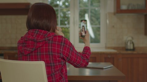 Back view of woman recording video using mobile phone vertically sitting in the kitchen. Female talking to mobile phone camera.