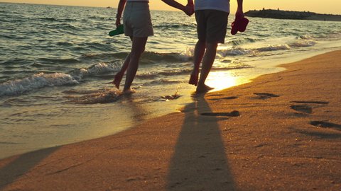 Couple walking together on the beach at sunset, young happy couple holding hands walking along beach, walking barefoot and carrying shoes, outdoor leisure time by the seaside.