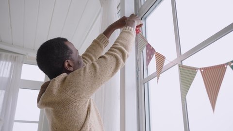 African American man hangs colorful patterned pennant banners on window. Happy black guy prepares for birthday party decorating room at home