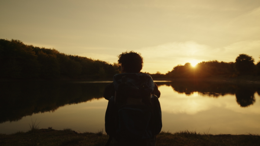 boy with backpack sits near the lake at sunset