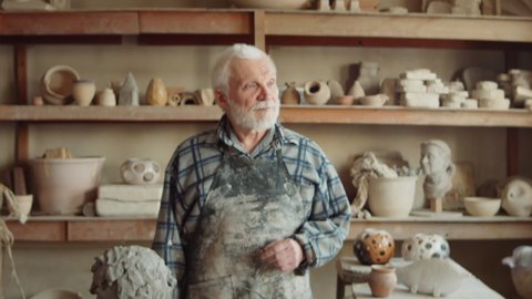 Zoom in portrait shot of senior potter with grey hair and beard standing in workshop and posing for camera