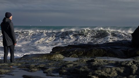Panning Slow Motion Wide Shot Of Man Standing On Rocky Shore And Moving To Dodge Crashing Waves, Hoffen, Iceland