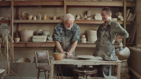 Zoom in shot of senior potter kneading clay and explaining something to young woman during ceramics lesson in workshop