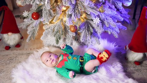 Caucasian child in green elf costume and red socks with deer resting on the fluffy plaid. Beautiful toddler moving legs actively under Christmas tree.