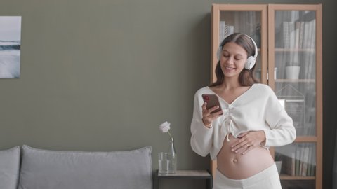 Medium slowmo shot of happy young pregnant woman with smartphone in hands dancing to music in headphones and stroking her belly