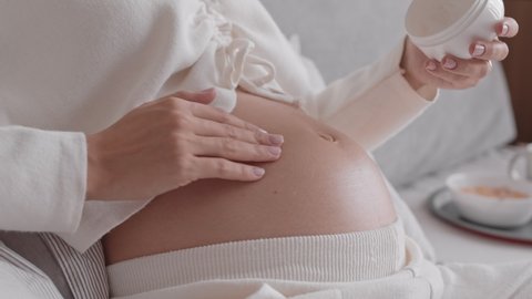 Slowmo closeup of unrecognizable pregnant woman rubbing moisturizing cream on her belly, taking care of her skin during pregnancy