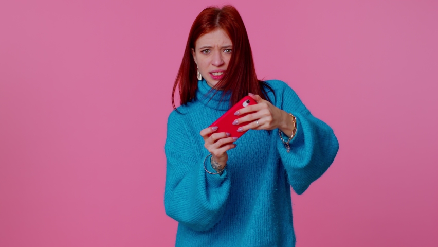 Worried funny addicted woman enthusiastically playing racing video games on mobile phone. Young adult teen girl using smartphone gadget app with drive simulator isolayed on pink studio wall background