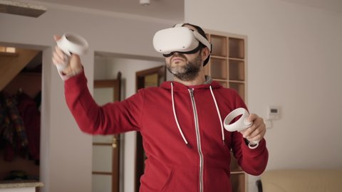 Man At Home Wearing Virtual Reality Headset Holding Gaming Controllers. VR Tester in White Headset Moves Hands With Controllers Man Playing Games On Virtual Reality Device. Metaverse Oculus Technology