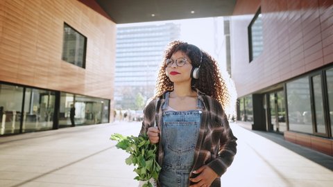Curly african american woman in eyeglasses and headphones is enjoying life and music while going home from the market with green celery in the bag outdoors.