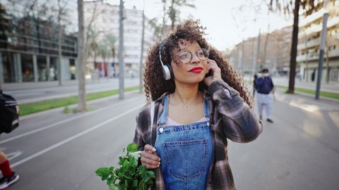 Outdoors portrait of african american woman in eyeglasses and headphones going home after shopping from the market with green celery in eco bag.