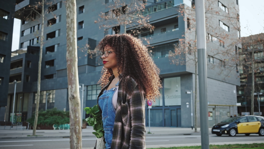 Side view of a beautiful long-haired mixer race girl in glasses and denim overalls walks through the city with a shopping bag with fresh greens on her shoulder. Vegetarianism and tourism concept.
