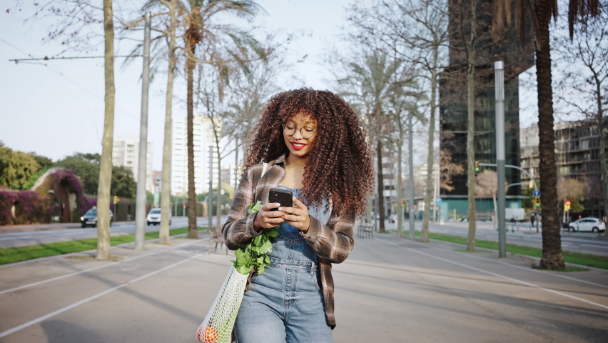 A girl wearing denim overalls and glasses walks along streets of barcelona with a phone in her hands. Happy youg tourist.
