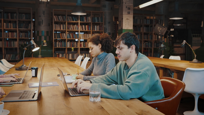 Concentrated diverse millennial students work at public library within small group of mates. Young Black female create ideas take notes on paper sheets while her male friend browse web on laptop pc