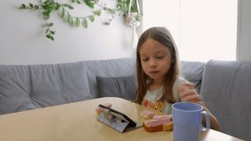 a little girl eats sausage sandwiches and watches cartoons on a smartphone screen, sitting at a table in the kitchen, home life