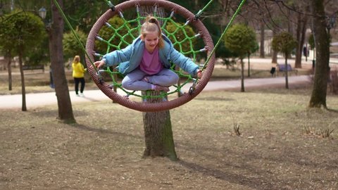 A cheerful little girl in a blue coat rides on a swing on the playground in park. walks with children. improvement of territories.