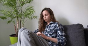 One girl with black curly hair and fair skin sits at home on a gray sofa and looks at the phone, there is a palm-like plant nearby, the walls in the room are white, her legs are bent