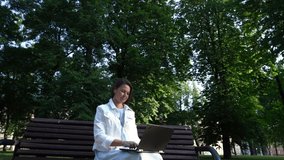 Businesswoman in white jacket greets colleague talking with partner via laptop sitting on shadow bench in park slow motion