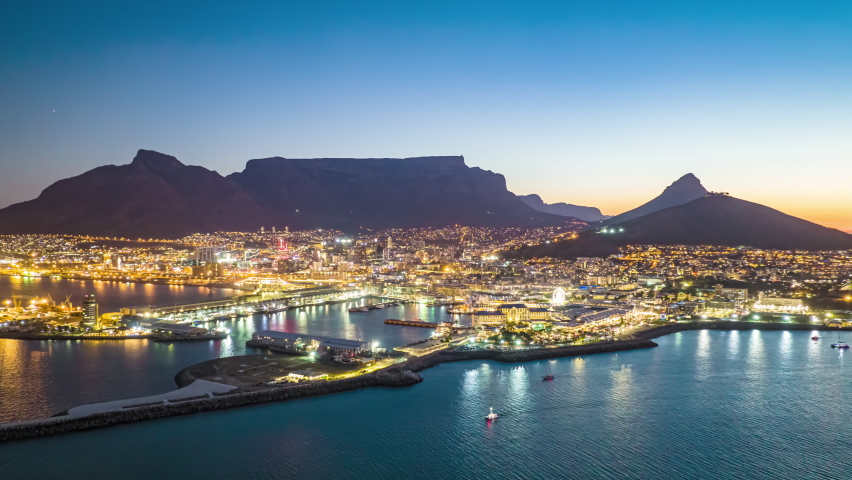 Aerial panoramic hyperlapse shot of city at dusk. Illuminated buildings and streets with Table mountain silhouette in background. Fly above marina. South Africa Royalty-Free Stock Footage #1089162575