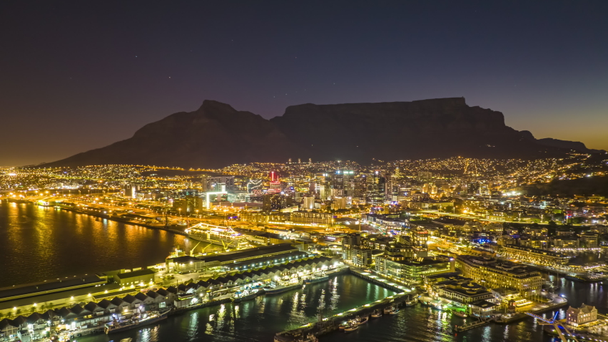 Aerial panoramic hyperlapse shot of city at dusk. Illuminated buildings and streets with Table mountain silhouette in background. Fly above marina. South Africa