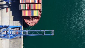 Aerial drone top down pan video above huge cranes load - unload colourful anchored container tanker in logistics terminal port