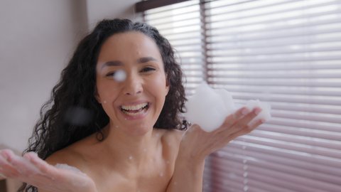 Close-up funny happy playful young naked cute woman playing in bathroom blowing foam smiling laughing fooling around relaxing in bathtub at home having fun having free time enjoying play with soap