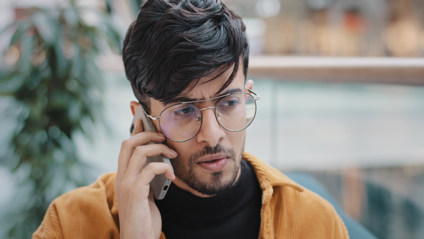 Portrait worried annoyed young arab man standing indoors talking on telephone answering call using smartphone guy angry bad cell service mobile connection problem low battery broken phone microphone Royalty-Free Stock Footage #1089164065