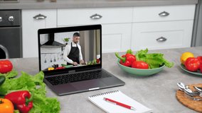 Laptop computer with man food blogger shows slice cucumber tells teaches housewife students stands on kitchen table near ingredients vegetables and notebook with pen, online video call cooking lesson