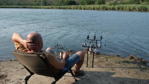Fisherman with hot drink in mug waiting a bite with carp fishing equipment. Angler is fishing with carpfishing technique in a beautiful summer evening, using rod pod, bite alarms, swinger, rods