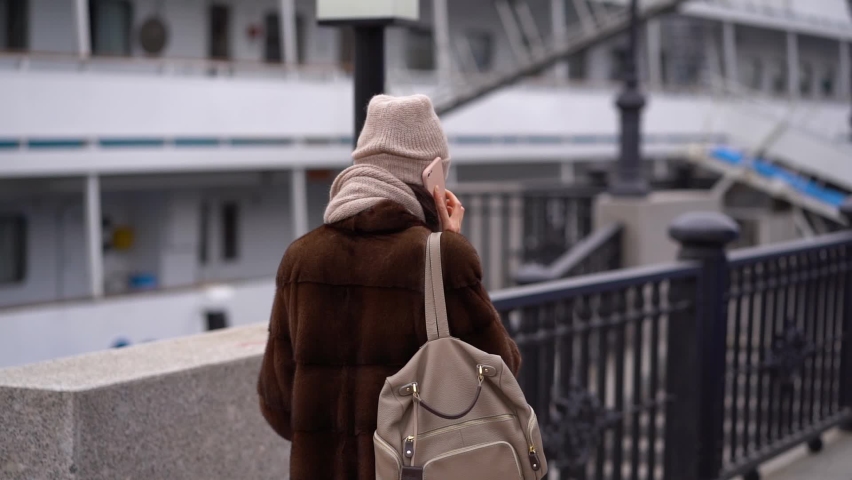 Back view of mature woman in winter clothes walking to home talking on phone. European girl using cellphone on city street with urban background. Slow motion.