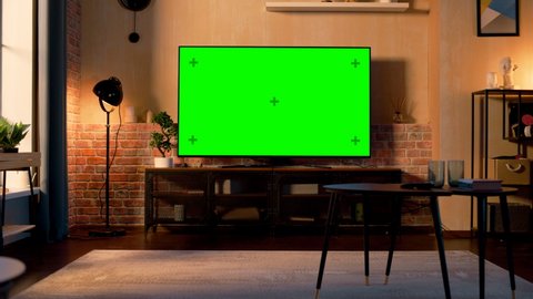 Stylish Loft Apartment Interior with TV Set with Green Screen Mock Up Display Standing on Television Stand. Empty Living Room at Home with Chroma Key Placeholder on Monitor. Zoom In Sunset Warm Shot.