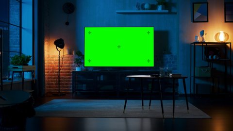 Stylish Loft Apartment Interior with TV Set with Green Screen Mock Up Display Standing on Television Stand. Empty Living Room at Home with Chroma Key Placeholder on Monitor. Zoom In Night Shot.
