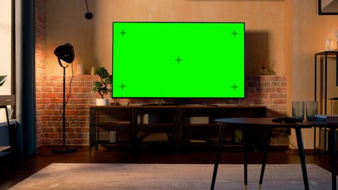 Stylish Loft Apartment Interior with TV Set with Green Screen Mock Up Display Standing on Television Stand. Empty Living Room at Home with Chroma Key Placeholder on Monitor. Zoom Out Sunset Warm Shot.