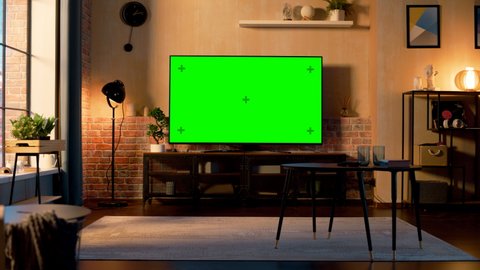 Stylish Loft Apartment Interior with TV Set with Green Screen Mock Up Display Standing on Television Stand. Empty Living Room at Home with Chroma Key Placeholder on Monitor. Still Sunset Warm Shot.